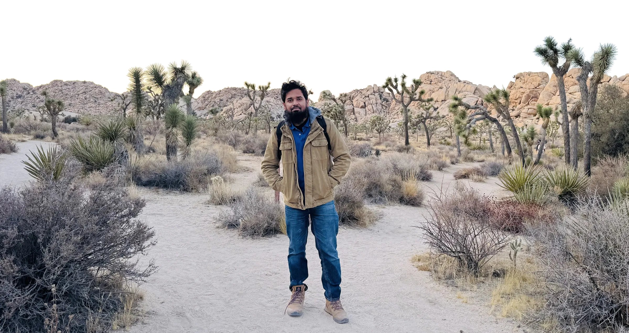 Affan on a sandy trail in Joshua Tree National Park at sunset, with Joshua trees and granite boulders behind him