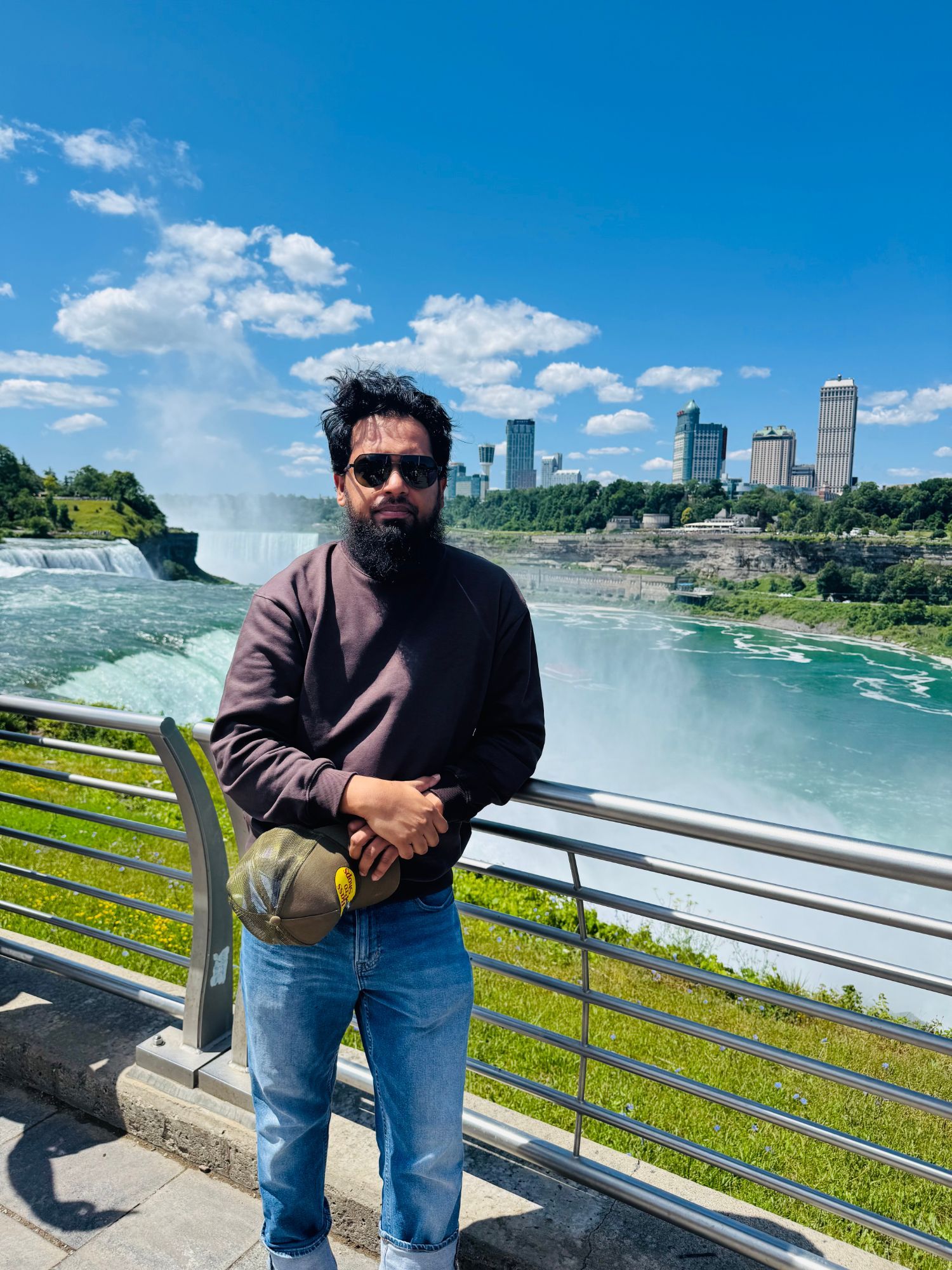 Affan in sunglasses leaning on a railing above Niagara Falls, with the river and the city skyline behind him